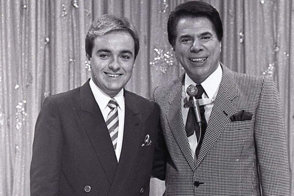 Foto antiga em preto e branco do apresentador Gugu e Silvio Santos, também apresentador e dono do SBT. Eles aparecem sorrindo e abraçados - Metrópoles