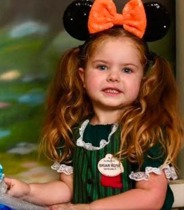 Foto colorida de uma menina branca e loira, vestindo um vestido verde e um laço laranjado no cabelo
