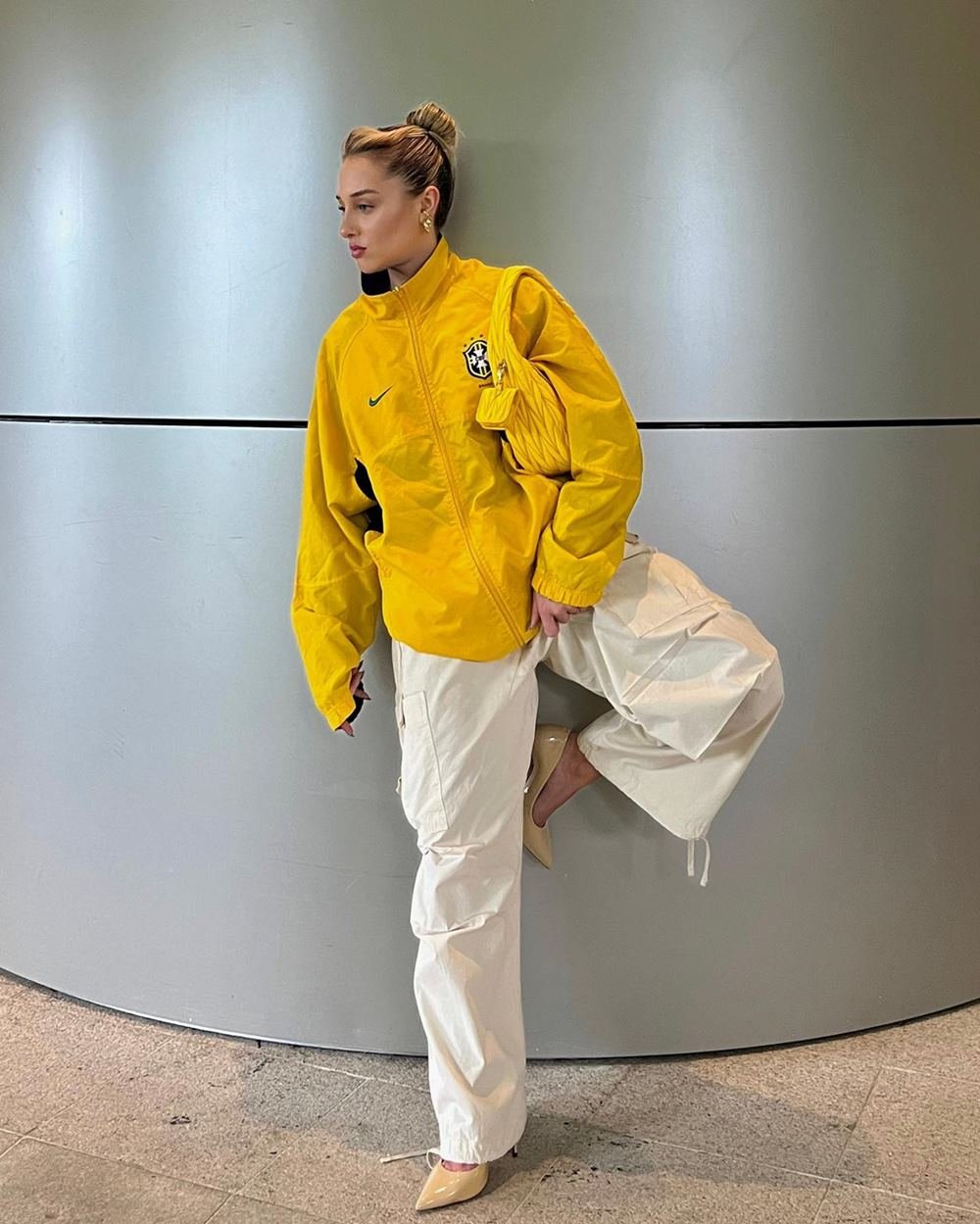 A influencer Malu Borges, uma mulher branca, jovem e loira, posando para foto em uma parede cinza. Ela usa uma calça folgada branca, um salto alto bege, uma jaqueta amarela com o símbolo da CBF e um bolsa, também amarela, da marca Miu Miu.