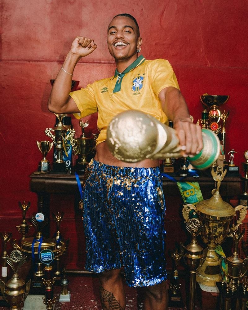 Homem jovem e negro, de cabelo curto e bigode, segurando uma réplica da taça da Copa do Mundo de Futebol. Ele usa um shorts azul de lantejoulas e uma camiseta de seleção brasileira.