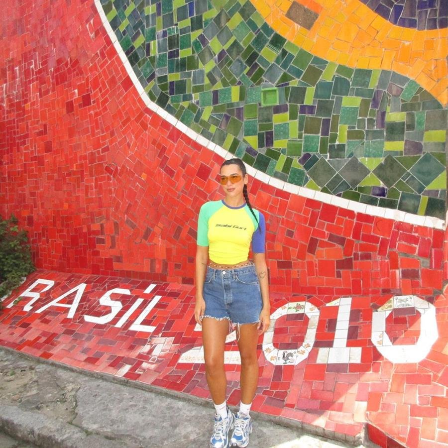 A cantora Dua Lipa, um mulher branca, jovem e de cabelo liso longo, posando para foto na Lapa, no Rio de Janeiro. Ela usa uma camiseta com as cores do Brasil, um shorts jeans e um tênis branco.