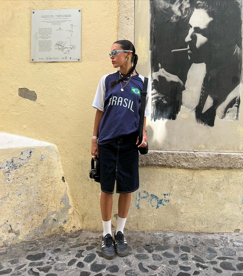 Uma mulher branca e jovem, com cabelo castanho liso amarrado em uma trança, nas ruas de Lisboa, em Portugal. Ela usa óculos escuros, uma camiseta da seleção brasileira de futebol na cor azul, um bermuda preta, meias brancas e um tênis preto da marca Adidas.