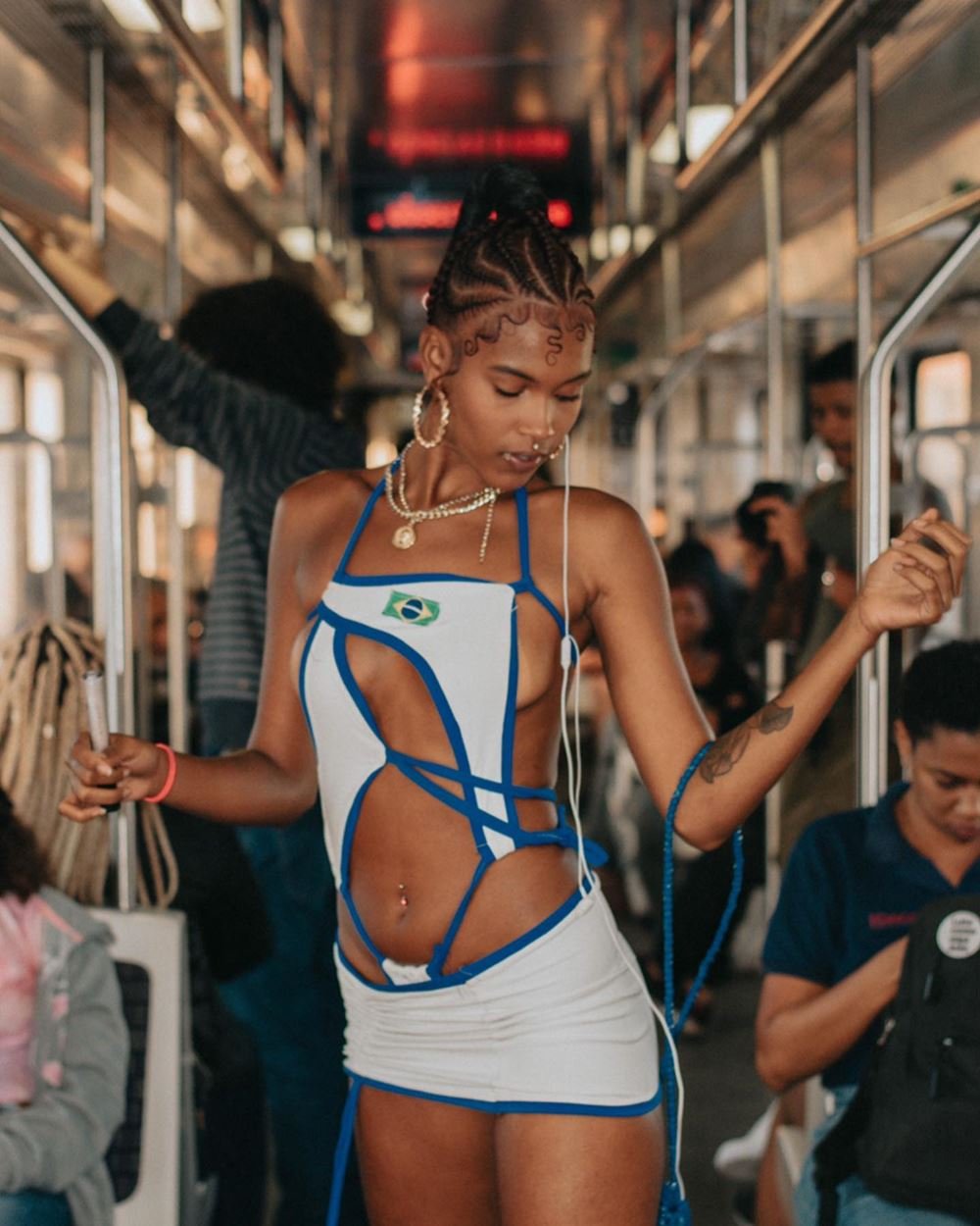 Mulher jovem e negra, de cabelo trançado longo, posando para foto em um vagão do BRT. Ela usa um vestido branco cheio de recortes com uma bandeira do Brasil bordada e recortes na região da cintura.