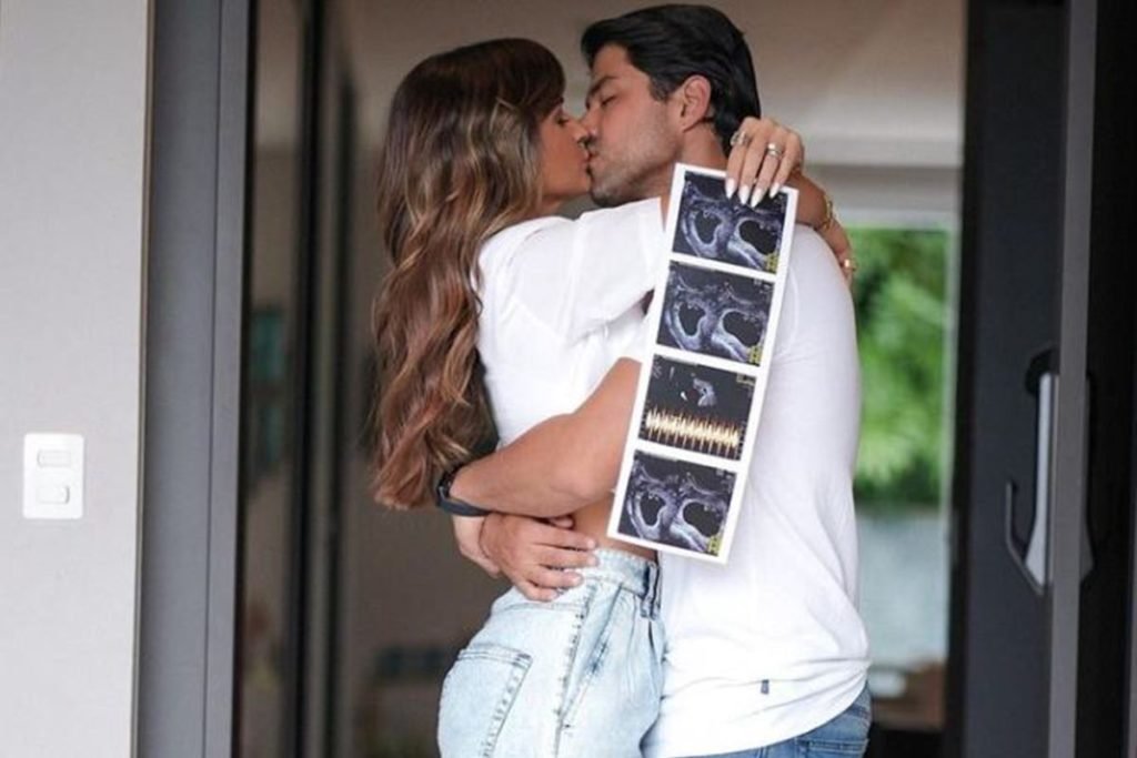 homem e mulher de blusa branca e jeans abraçados se beijando segurando ecografias