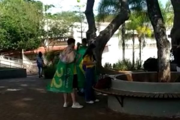 Pessoas com bandeira verde e roupa amarela em praça com árvores