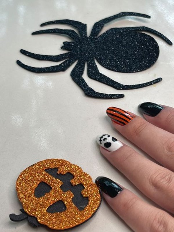 Foto colorida de quatro dedos com as unhas decorada e dois desenhos, uma aranha e uma abóbora de halloween