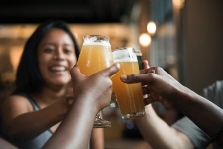 Foto com várias pessoas segurando copos com cerveja e brindando, Metrópoles.
