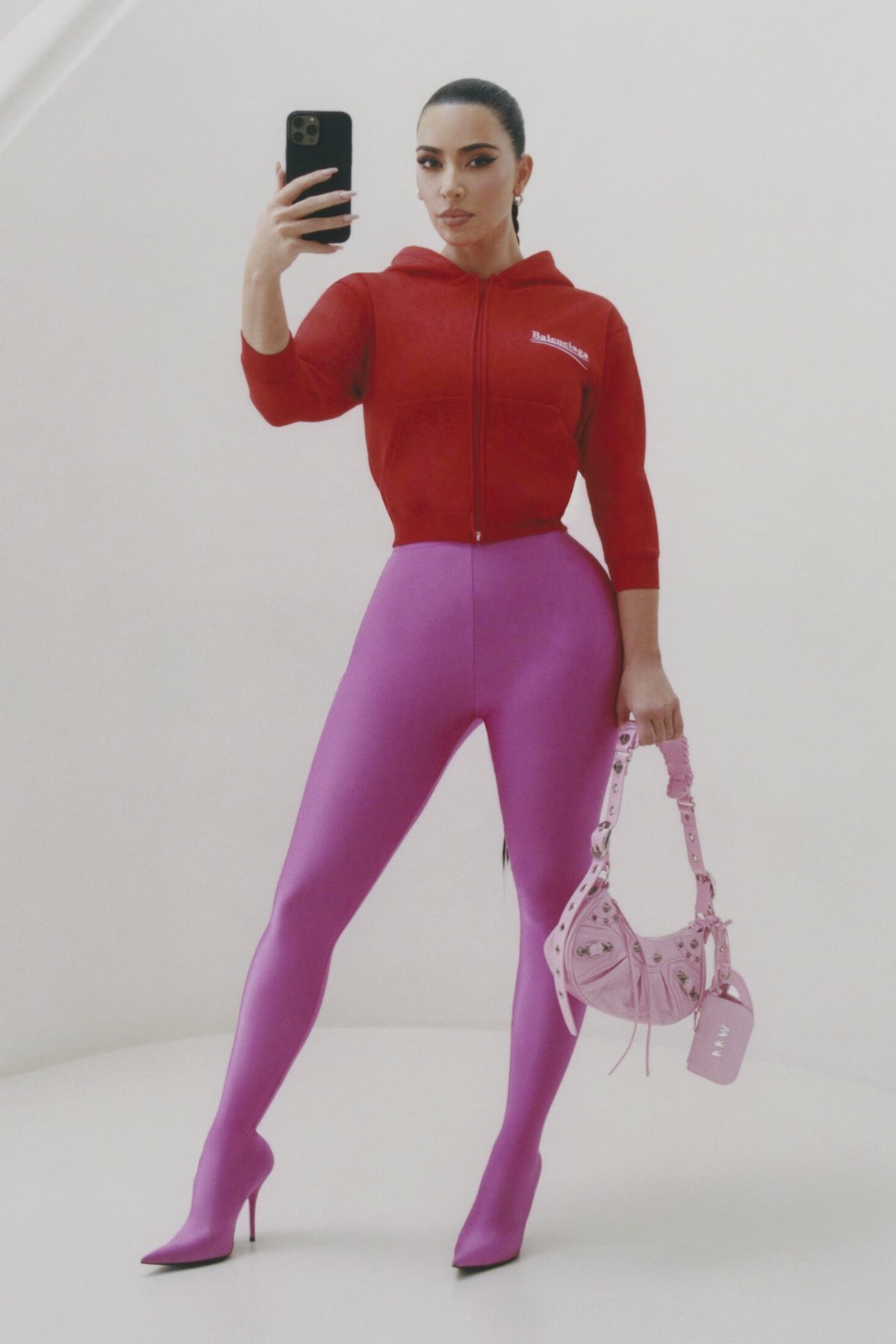 A empresária Kim Kardashian, uma mulher branca, de cabelo liso longo amarrado em uma trança, posa para a campanha da grife de luxo Balenciaga. Ela usa casaco de moletom vermelho, uma legging rosa escura e segura uma bolsa de couro rosa claro. - Metrópoles