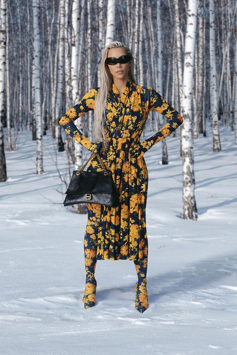 Foto da campanha de outono 2022 da grife de luxo Balenciaga. Na imagem é possível ver a empresária Kim Kardashian, uma mulher branca e jovem, com cabelo liso loiro, em uma floresta coberta por neve. Ela usa um vestido e uma bota, ambos com uma estampa de flores amarelas, uma bolsa preta de couro e óculos escuros pretos. - Metrópoles