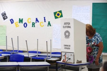 Mulher de cabelo ruivo em cabina eleitoral, na Escola Classe 7 de Ceilândia. Ao fundo, cadeiras escolares sobre carteiras. Na parede, lê-se a palavra "democracia", nas cores da bandeira do Brasil. Também há cartazes e uma bandeira nacional no mural - Metrópoles