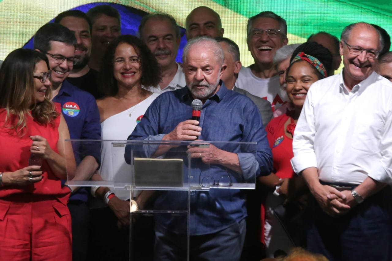 O presidente eleito Lula discursa cercado de aliados e do vice-presidente Geraldo Alckmin após vitória no segundo turno. Ao fundo, um painel com a bandeira do Brasil - Metrópoles