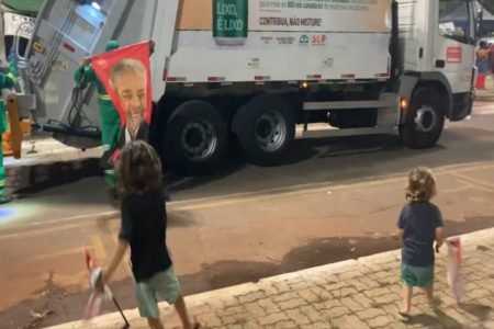 Foto de duas crianças e um gari com uma bandeira em frente a um caminhão de lixo
