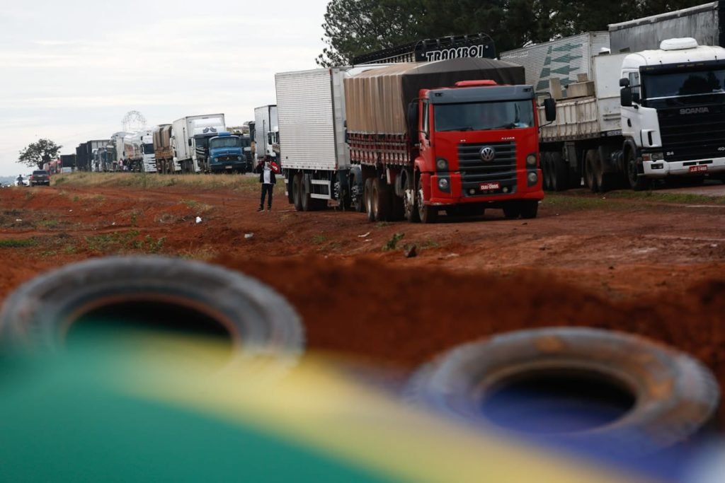Caminhoneiros e pessoas ligadas ao agro fecham a BR-251 no quilômetro 14, liberando somente serviços de saúde. Na imagem, caminhões parados e pneus jogados pela terra aparecem atrás de bandeira do Brasil - Metrópoles