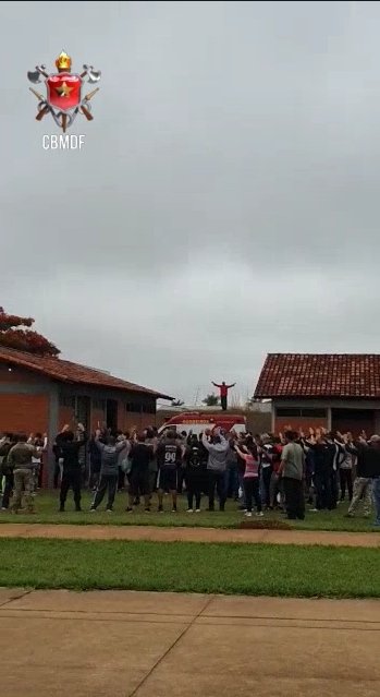 Foto colorida. Multidão de costas entre duas casas. Homem de camisa vermelha em cima de uma ambulância com os braços abertos
