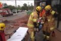Bombeiros militares levam homem imobilizado em prancha até maca. No fundo, um prédio em obra. Foto colorida