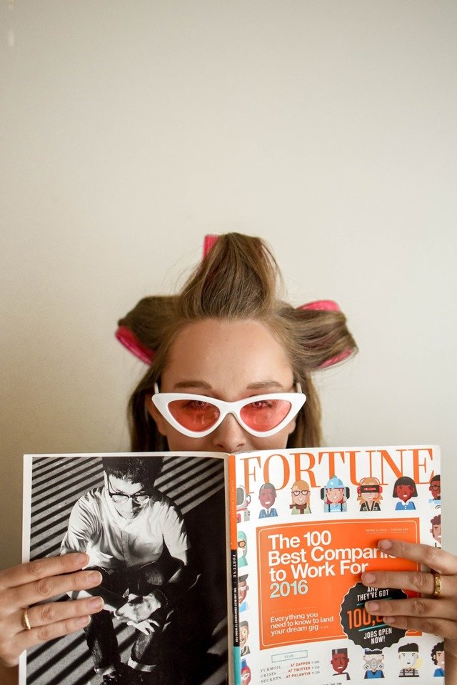 Jovem mulher de óculos com bobs no cabelo segurando revista - Metrópoles