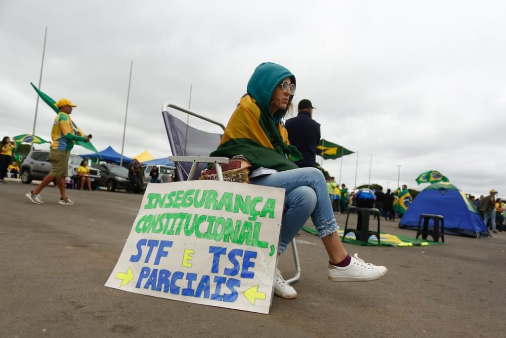 Manifestantes pró-Bolsonaro abraçada em bandeira do Brasil e com um cartaz com dizeres Insegurança institucional STF e TSE parciais, em frente ao Quartel General do Exército Forte Caxias, QG do exército em Brasília - Metrópoles