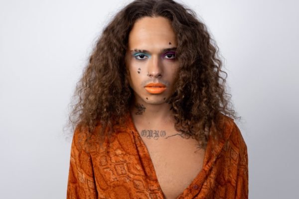 Foto colorida do cantor Vitão, cotado no BBB23, com maquiagem, batom e uma blusa laranja decotada - Metrópoles