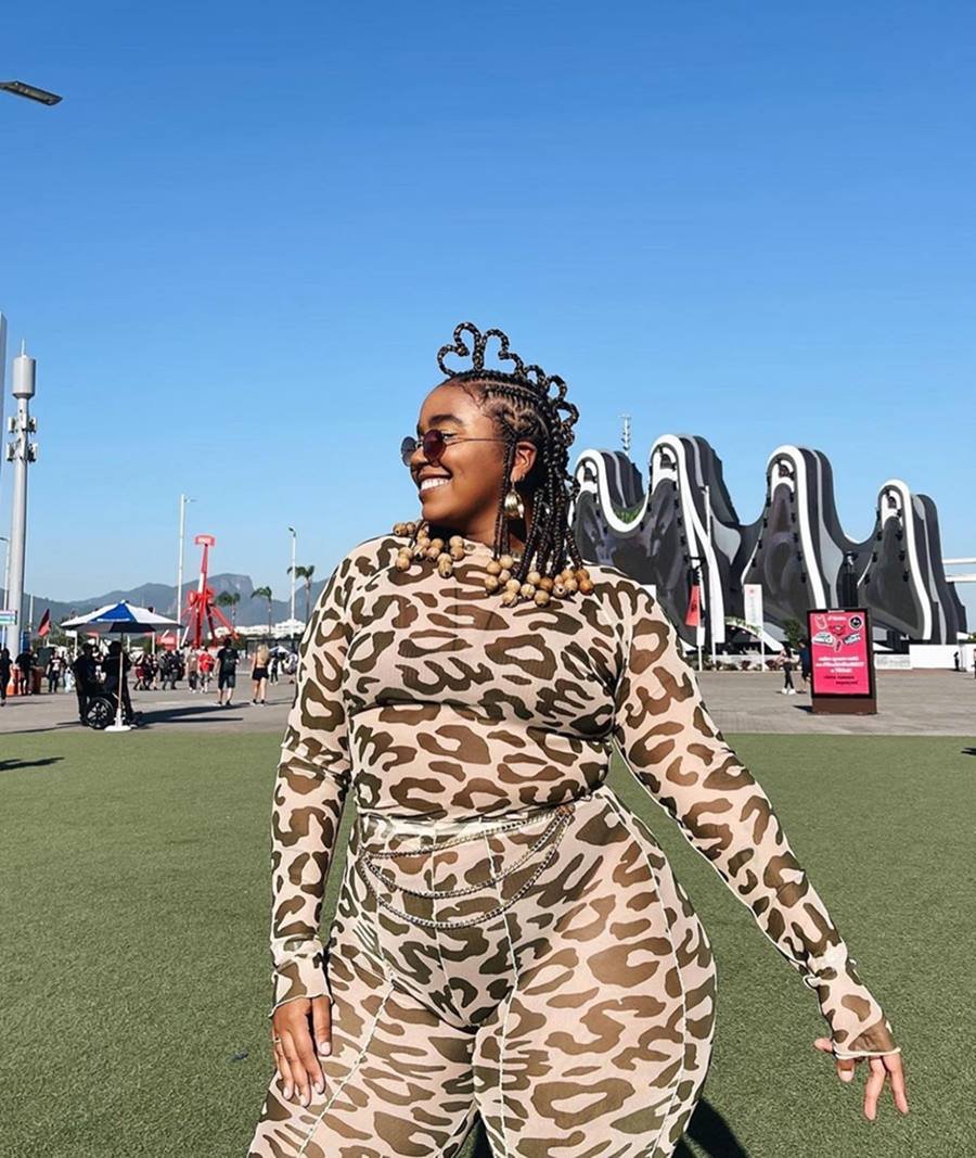 Mulher negra e jovem, de cabelo trançado, posando para foto no festival Rock in Rio. Ela usa um conjunto de blusa e calça com estampa de onça e óculos escuros. - Metrópoles