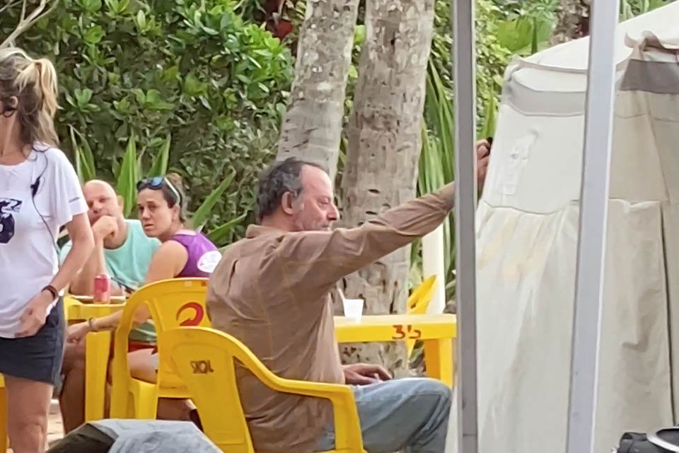 Foto colorida do ator Jean Reno vestindo calça jeans e camisa marrom sentado em uma cadeira amarela de bar no set de gravações de novo filme em Ubatuba, SP - Metrópoles