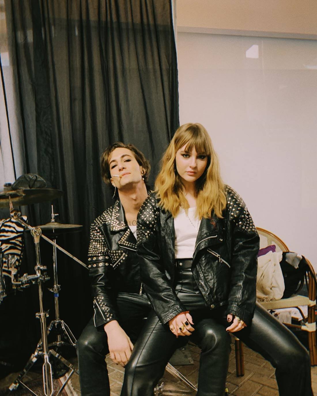 Mulher branca e jovem, de cabelo liso loiro, posando para foto sentada no colo de um homem branco, jovem, de cabelo preto liso grande. Eles são da banda de rock Måneskin. Os dois usam looks iguais: camisa branca e calça + jaqueta de couro preto. - Metrópoles