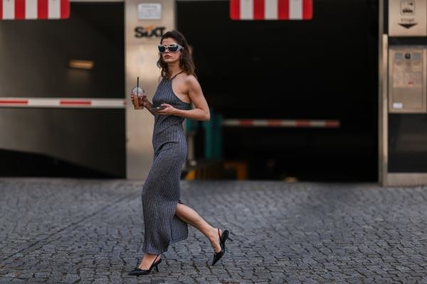 Mulher branca andando pela calçada. Ela está usando um vestido longo e óculos escuros. Também segura um café nas mãos - Metrópoles