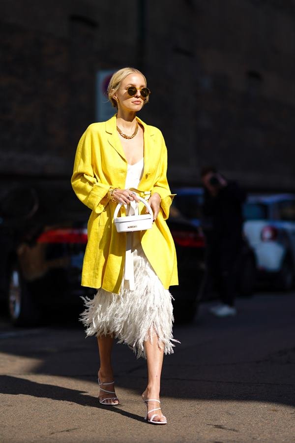 Mulher branca andando pelas ruas com vestido longo e um blazer amarelo. Ela está com óculos escuros e está segurando uma bolsa nas mãos - Metópoles