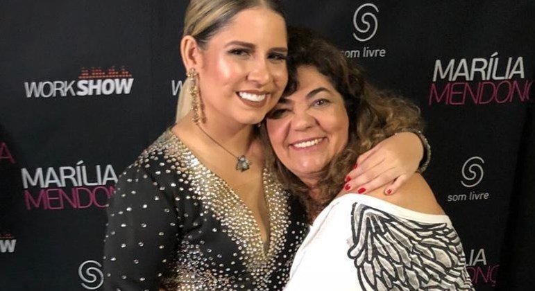 Na foto, a ex-assessora Silvia Colmenero e a cantora Marília Mendonça se abraçam em um fundo preto - Metrópoles