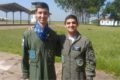 Homens brancos vestidos com casacos da Força Aérea verde colocam a mão no ombro um do outro. Eles olham para a câmera. Um deles usa lenço azul. Foto colorida