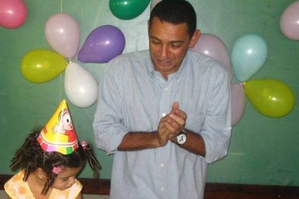 Fotografia colorida de homem em festa de criança cantando parabéns para menina ao lado dele