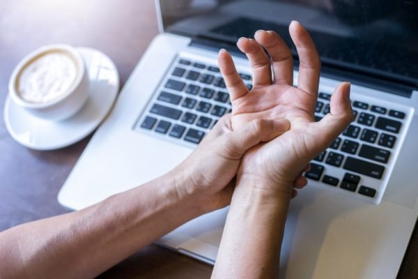 Pessoa apertando a mão com dor em frente a um teclado - Metrópoles