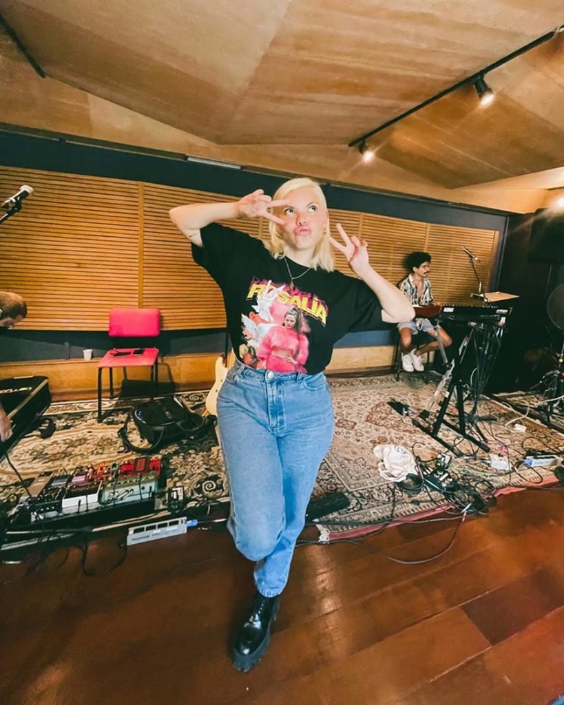 A cantora Duda Beat, uma mulher branca, jovem e com cabelo liso loiro, posando para foto em um estúdio de música.  Usa calça jeans, bota preta de couro e uma camiseta preta com estampa da cantora Rosalía.  - Metrópoles