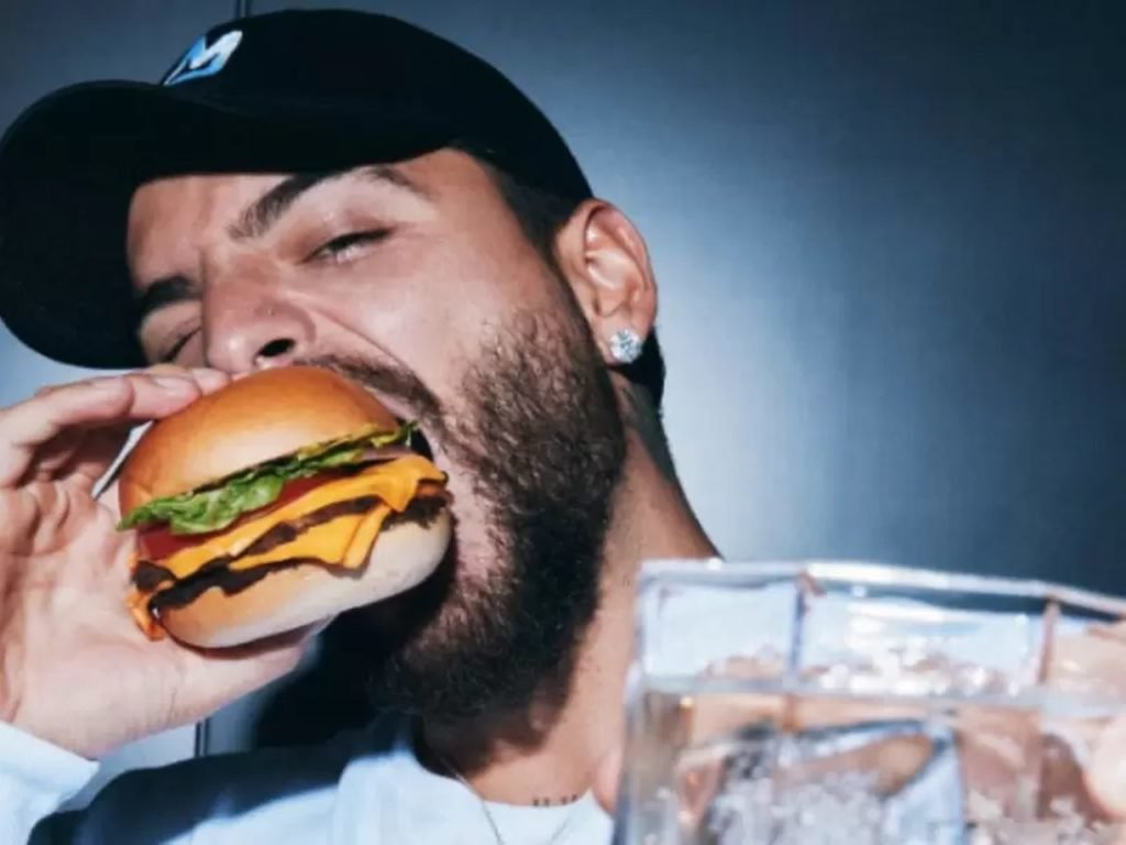 Na foto, o cantor Maluma posa para a câmera com um hambúrguer na boca - Metrópoles
