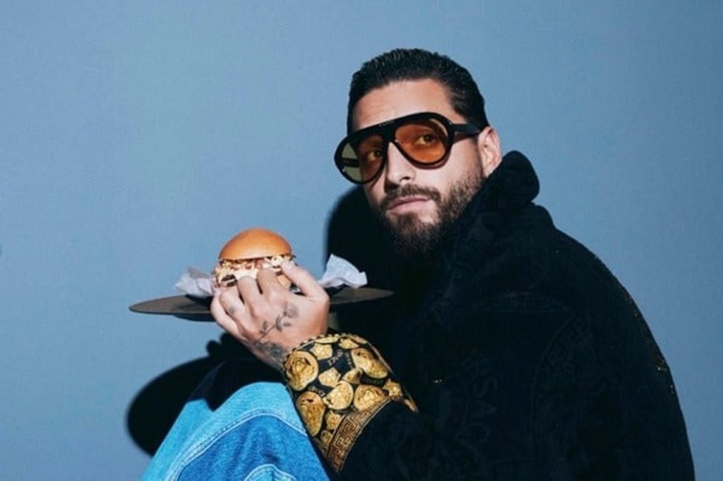 Na foto, o cantor Maluma posa para a câmera com um hambúrguer na mão - Metrópoles