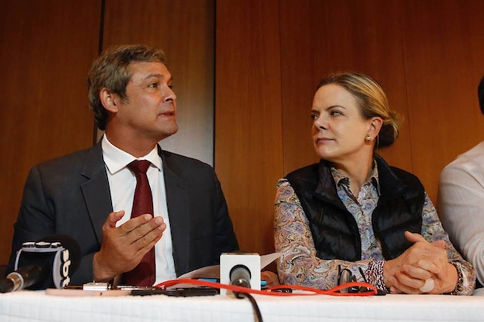 Os petistas Lindbergh Farias e Gleisi Hoffman durante evento. Enquanto Lindbergh fala, Gleisi o escuta, observando-o. Em sua frente, microfones - Metrópoles