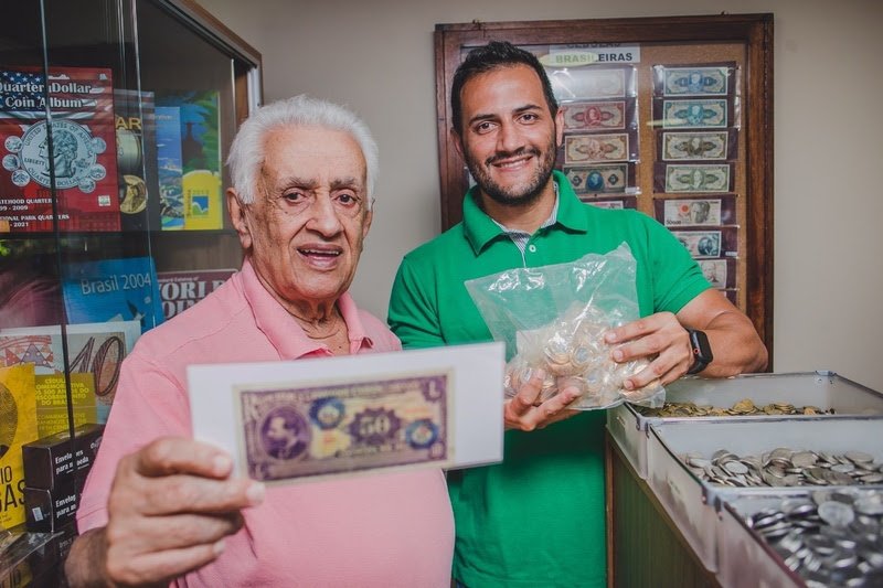 Homem idoso à frente da foto mostrando uma cédula antiga de dinheiro, ao lado um homem adulto de camisa verde segurando um pacote de moedas antigas