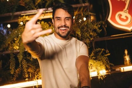 O DJ está usando camisa branca, está sorrindo e faz o simbolo do rock n roll - Metrópoles