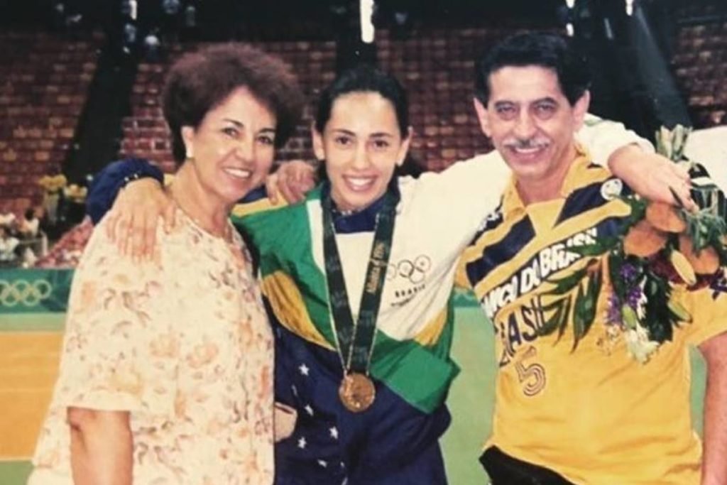 foto antiga de menina abraçada em um homem e uma mulher. Ela está ao centro e veste uniforme do brasil com medalha no pescoço - Metrópoles