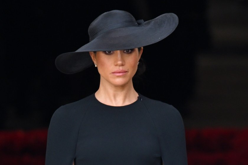Meghan Markle está usando roupa e chapéu preto. Sua expressão é de tristeza - Metrópoles