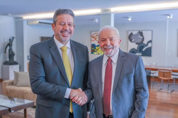 O presidente da Câmara dos Deputados, Arthur Lira, aperta a mão do presidente eleito Lula. Ambos olham para a câmera e sorriem, em sala - Metrópoles