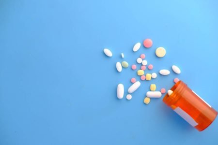Imagem ilustrativa de comprimidos diversos espalhados sob fundo azul e pote de remédio laranja e cilíndrico caído - Metrópoles