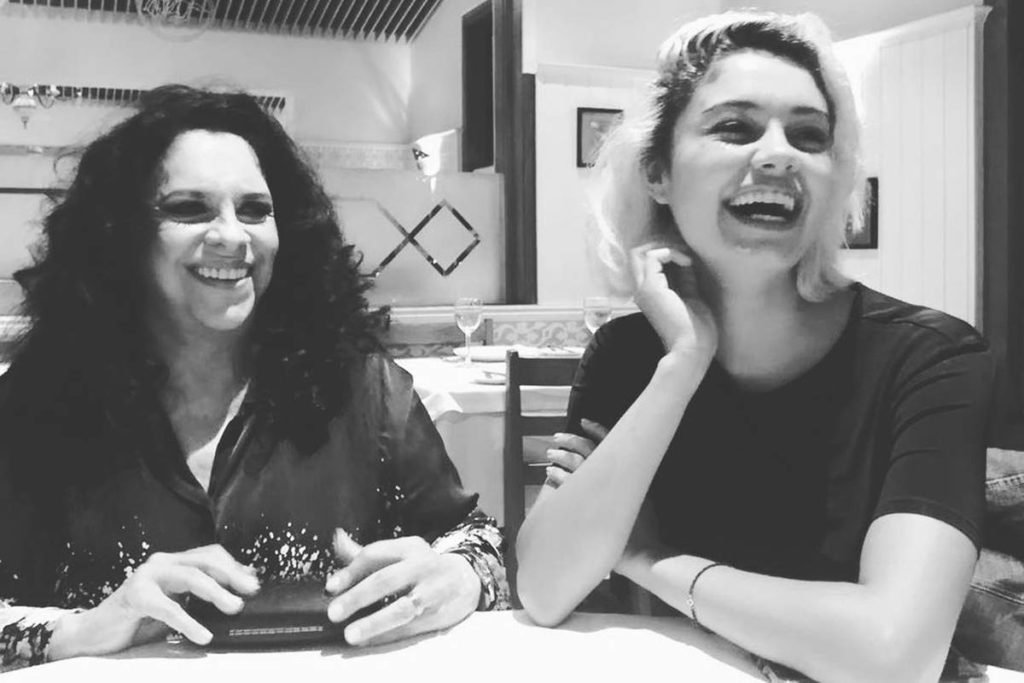 mulher de cabelos cacheados escuros sentada ao lado de mulher de cabelos curtos loiros sorrindo em foto preto e branco - Metrópoles