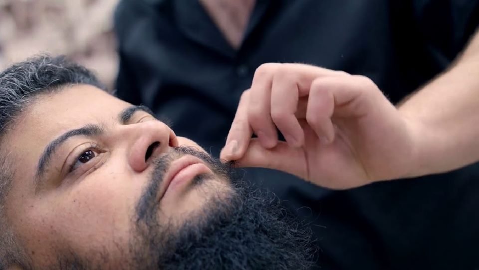 Homem de pele clara, cabelo preto e barba grande e preta sendo tocado por uma mão de pele clara - Metrópoles