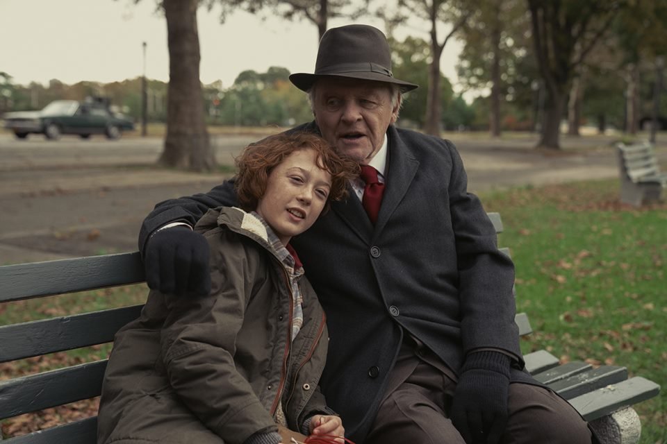 Garota com cabelo ruivo curto, pele clara, jaqueta escura com detalhes vemelhos abraçada por um homem velho, de chapéu marrom, cabelos brancos, pele clara, terno escuro, gravata vermelha, luvas pretas. Os dois estão sentados em um banco, em um parque - Metrópoles
