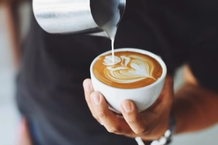 Na foto, uma mão segura uma xícara branca enquanto despeja leite vaporizado sobre o café - Metrópoles