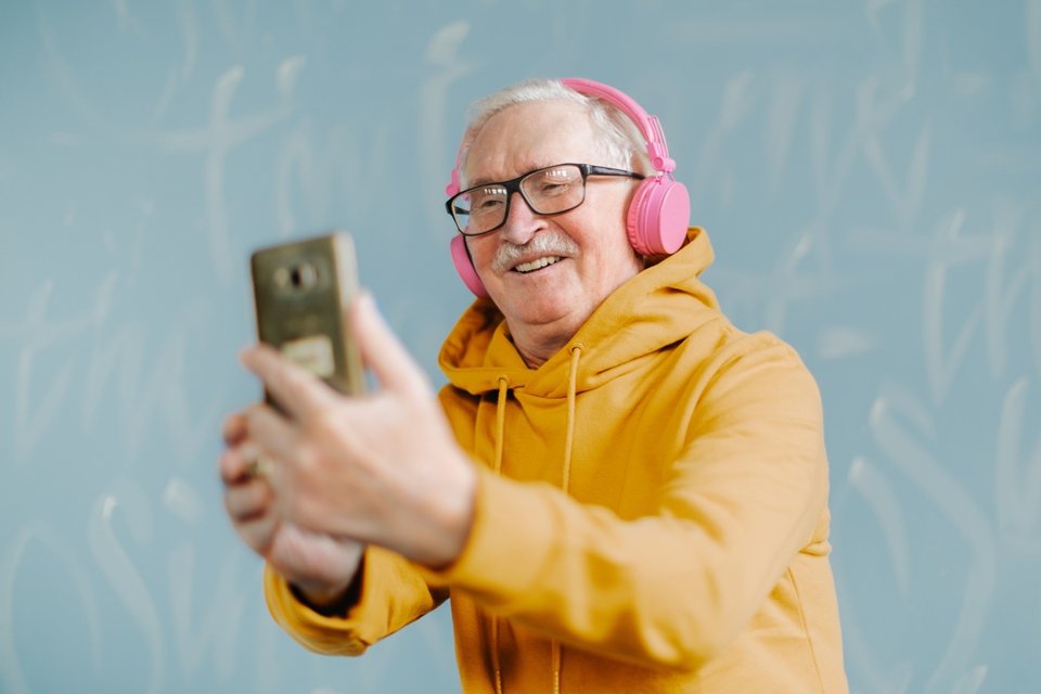 Foto colorida de home idoso com casaco amarelo, fones cor-de-rosa e segurando um celular - Metrópoles