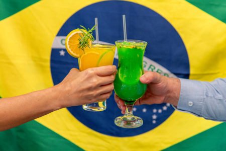 Na foto, duas pessoas brindando com drinques verde e amarelo com a bandeira do Brasil atrás - Metrópoles