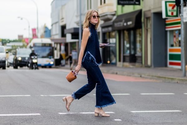 Mulher jovem, branca e loira, de cabelo liso, andando pelas ruas de Sydney, na Austrália. Usa uma regata jeans longa, uma calça jeans wide lege e uma sandália de salto baixo dourado. - Metrópoles