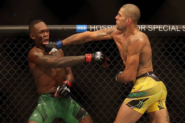 Em imagem colorida, o brasileiro Alex Poatan está de calção amarelo e dá um soco no nigeriano Israel Adesanya, de calção verde, no UFC 281 - Metrópoles