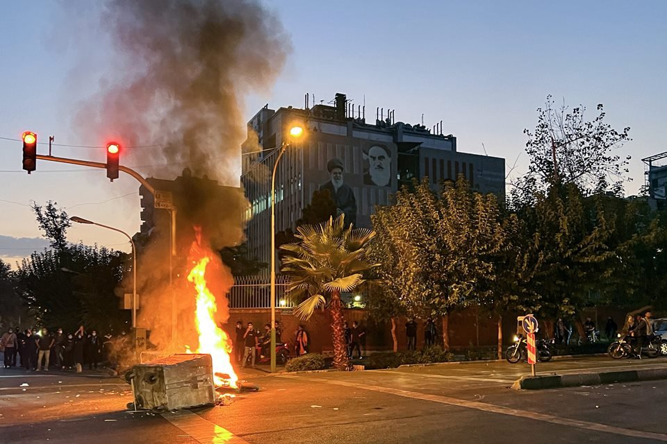 Em imagem colorida, lata de lixo pega fogo em uma rua de Teerã, no Irã, com imagens de líderes religiosos em um prédio ao fundo - Metrópoles